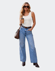 A woman stands on a wooden boardwalk beside a brick building, wearing a white ribbed tank top, light blue wide-leg jeans, a brown belt, and dark brown ankle boots. She holds a coffee cup and carries a brown studded shoulder bag.