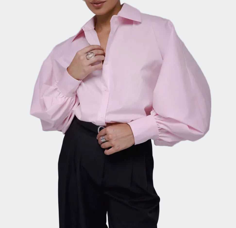A model wearing a light pink long-sleeve button-up blouse with dramatic balloon sleeves and black trousers, shown from the chin down against a plain light gray studio background.