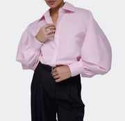 A model wearing a light pink long-sleeve button-up blouse with dramatic balloon sleeves and black trousers, shown from the chin down against a plain light gray studio background.