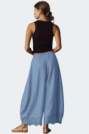 A woman shown from the back wearing a black sleeveless top and light blue wide-leg pants with lace trim at the hem, standing against a dark gray studio background.