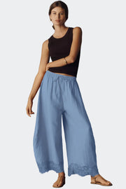 A woman modeling wide-leg light blue pants with a lace hem, paired with a black tank top and tan sandals, against a dark gray studio background.