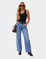 A woman stands on a wooden boardwalk beside a brick building, wearing a black sleeveless top, light blue wide-leg jeans, a brown belt, and dark brown ankle boots. She holds a coffee cup and carries a brown patterned shoulder bag.